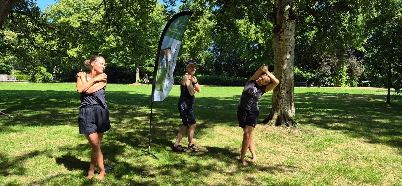 Drie deelnemers doen Fascial Maneuvers-oefeningen op het gras in het Rengerspark, met een Fit Natuerlik-vlag naast hen